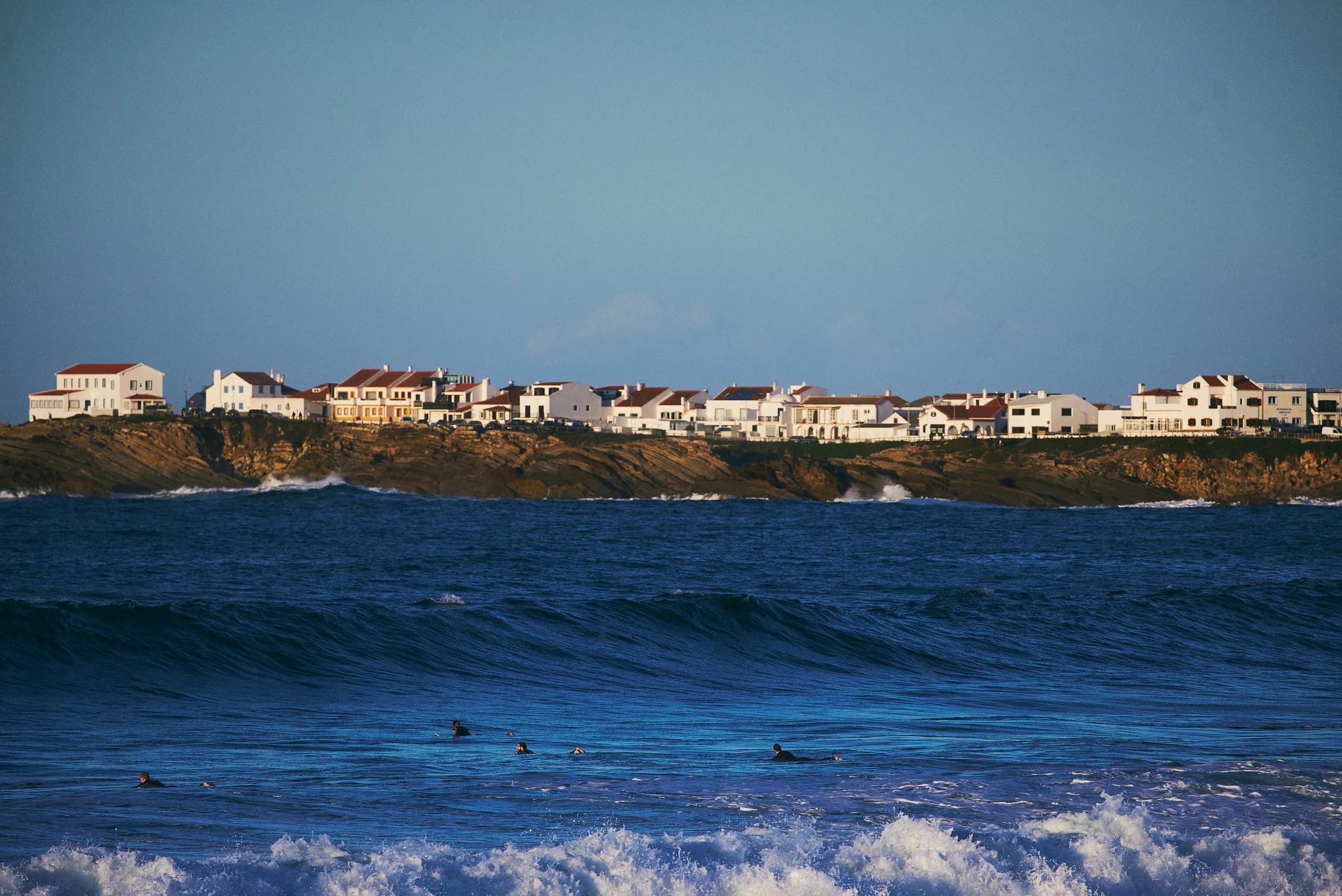 Surfistas esperando la ola frente a un pueblo costero de casas blancas