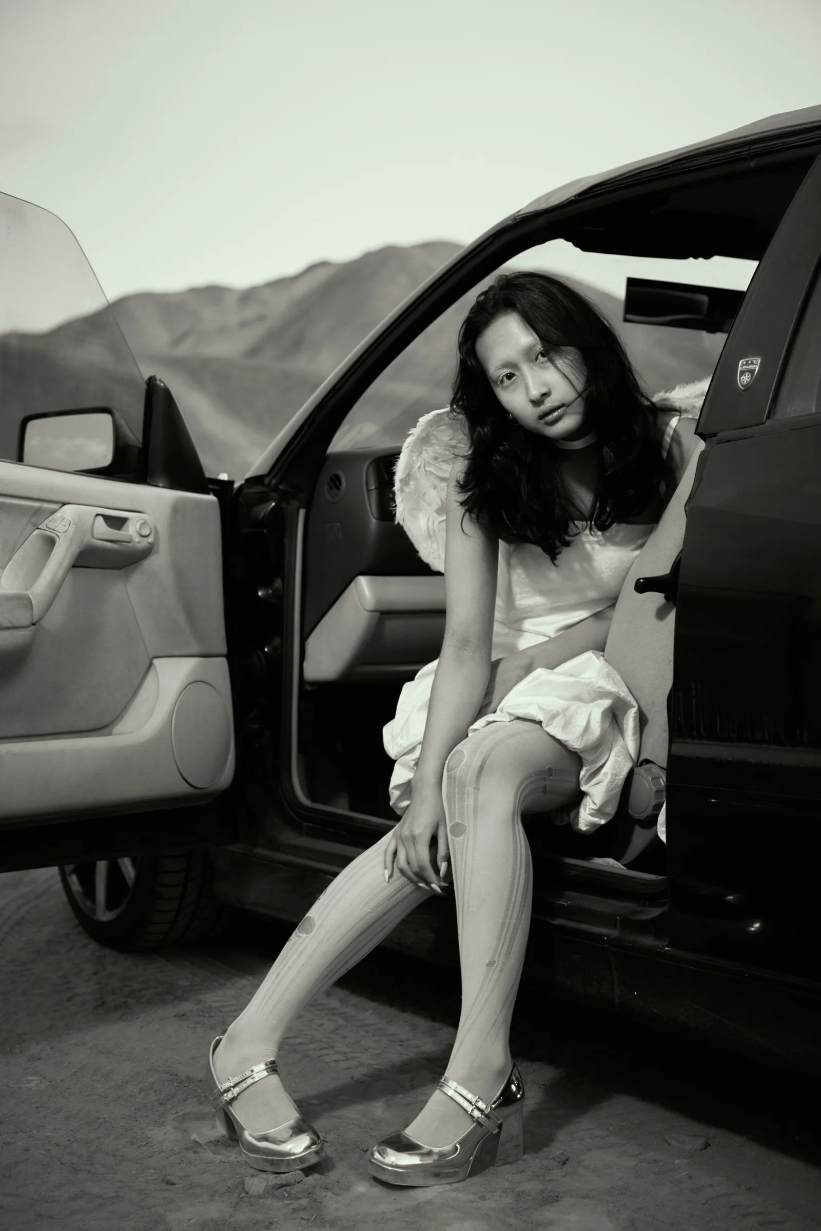 Retrato en blanco y negro de mujer con alas de ángel sentada en un auto en el desierto