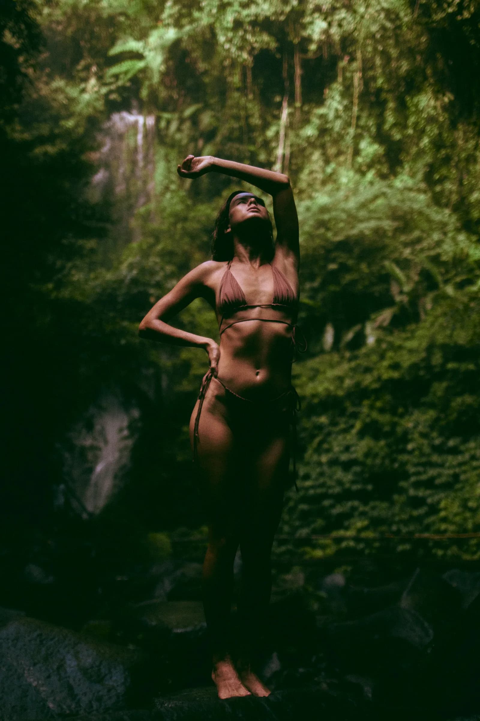 Mujer con traje de baño y el brazo en alto frente a una cascada en la selva