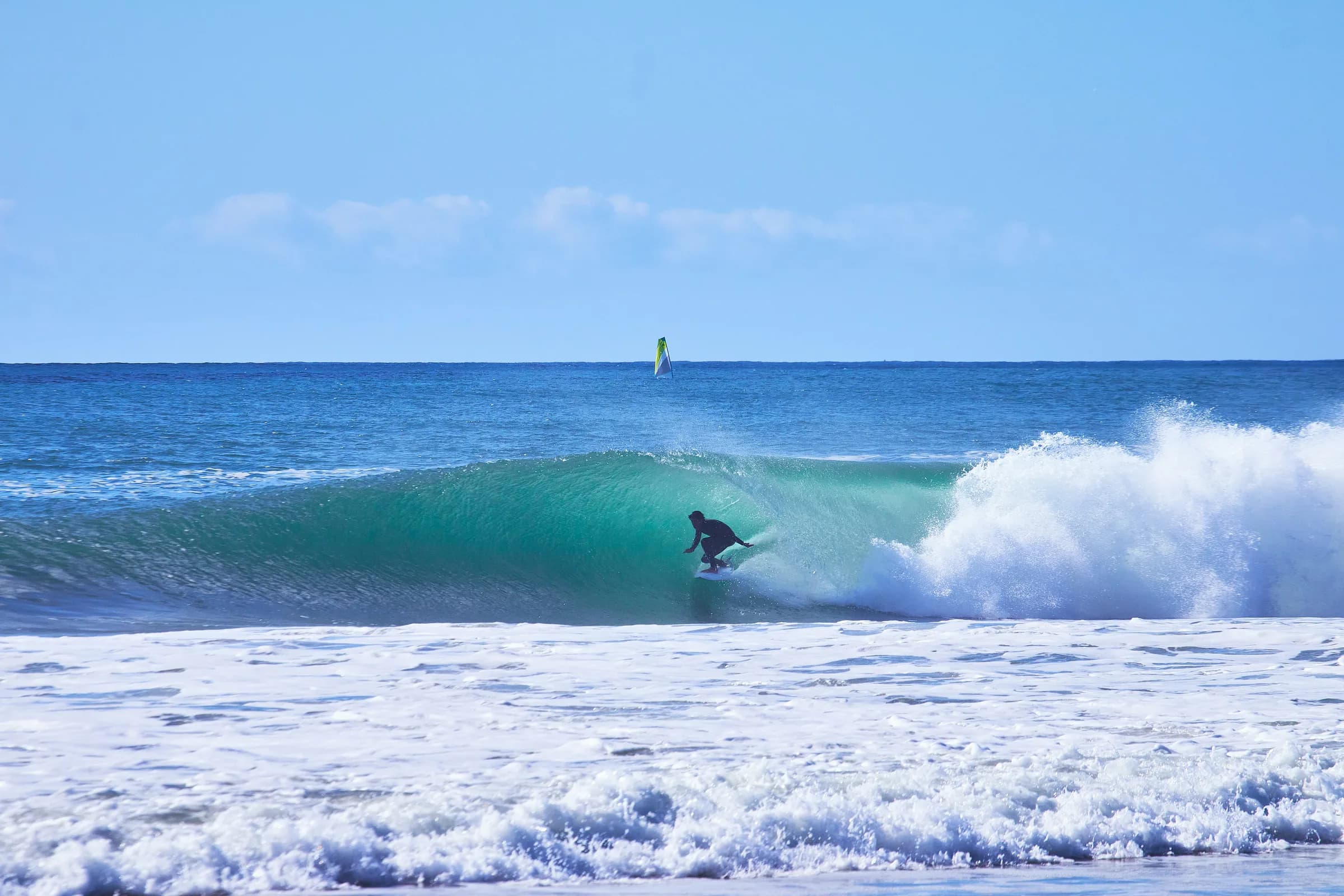 Surfista en el tubo de una ola turquesa con un windsurf al fondo